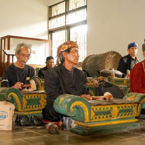 Elderly musicians playing traditional gamelan instruments indoors with regional attire.