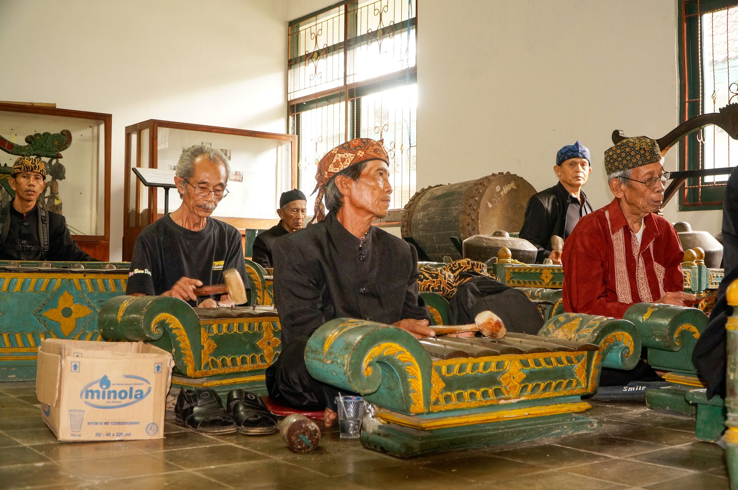 Elderly musicians playing traditional gamelan instruments indoors with regional attire.
